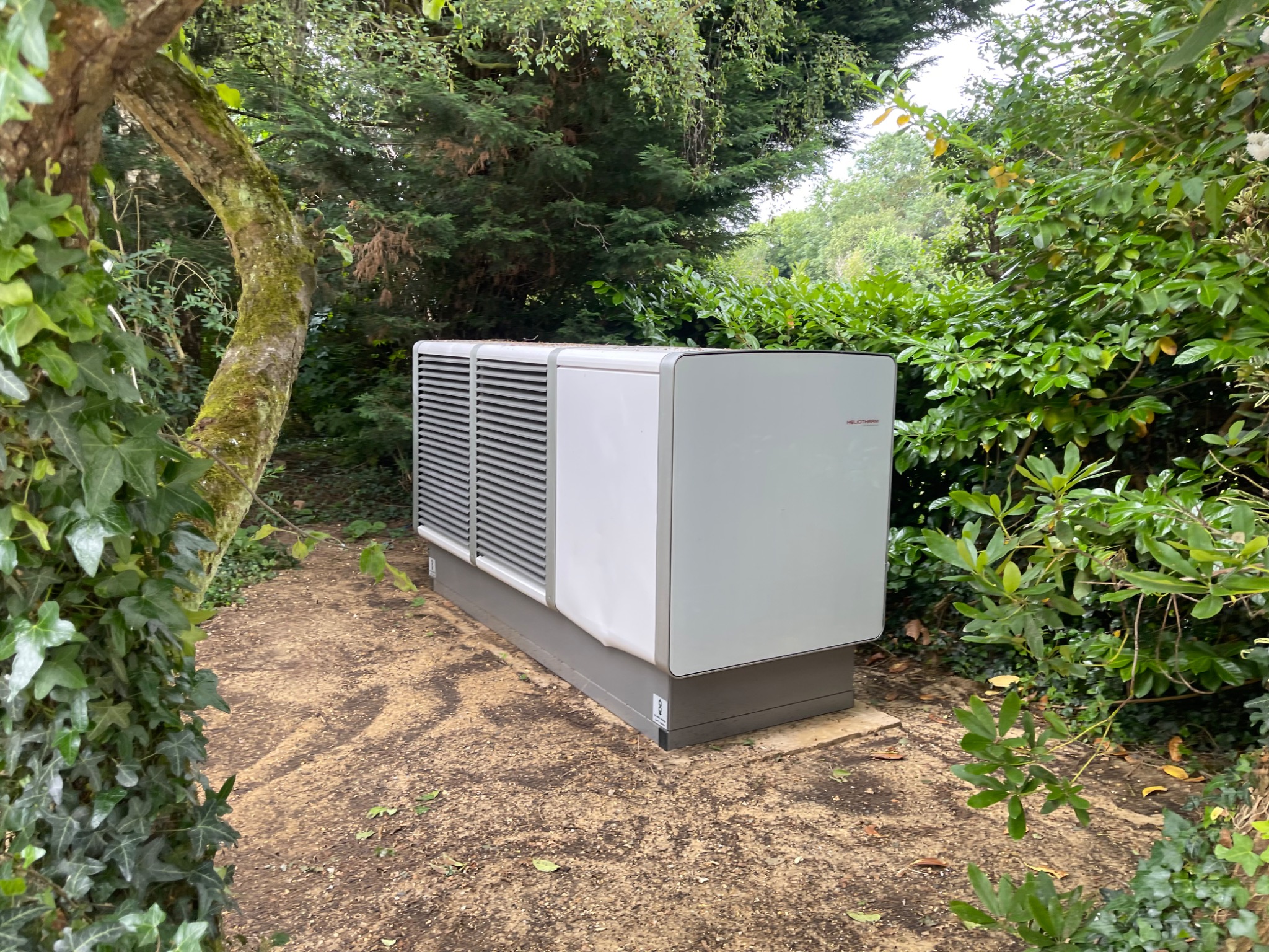 Heliotherm air source heat pump discreetly positioned within mature landscaping.