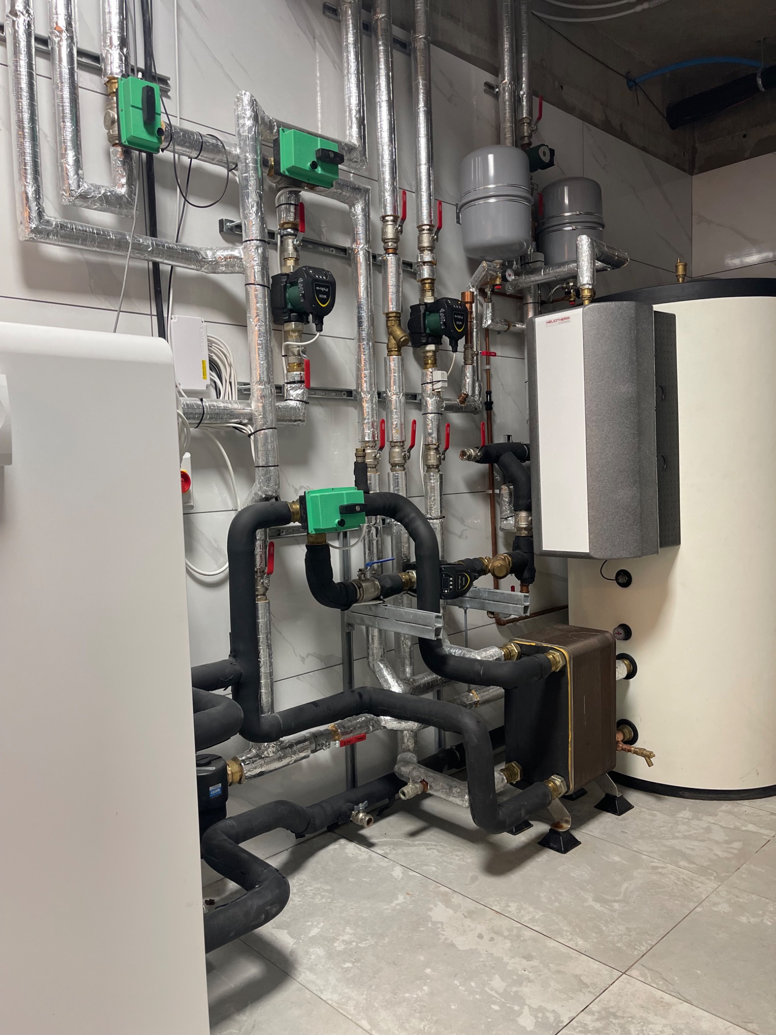 Plant room with black insulated brine pipework, plate heat exchanger and Heliotherm unit